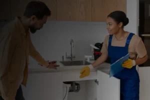Female plumber from Plumber Bradford Pros discussing a kitchen sink issue with a male homeowner, wearing work overalls and gloves, inspecting plumbing under the sink.